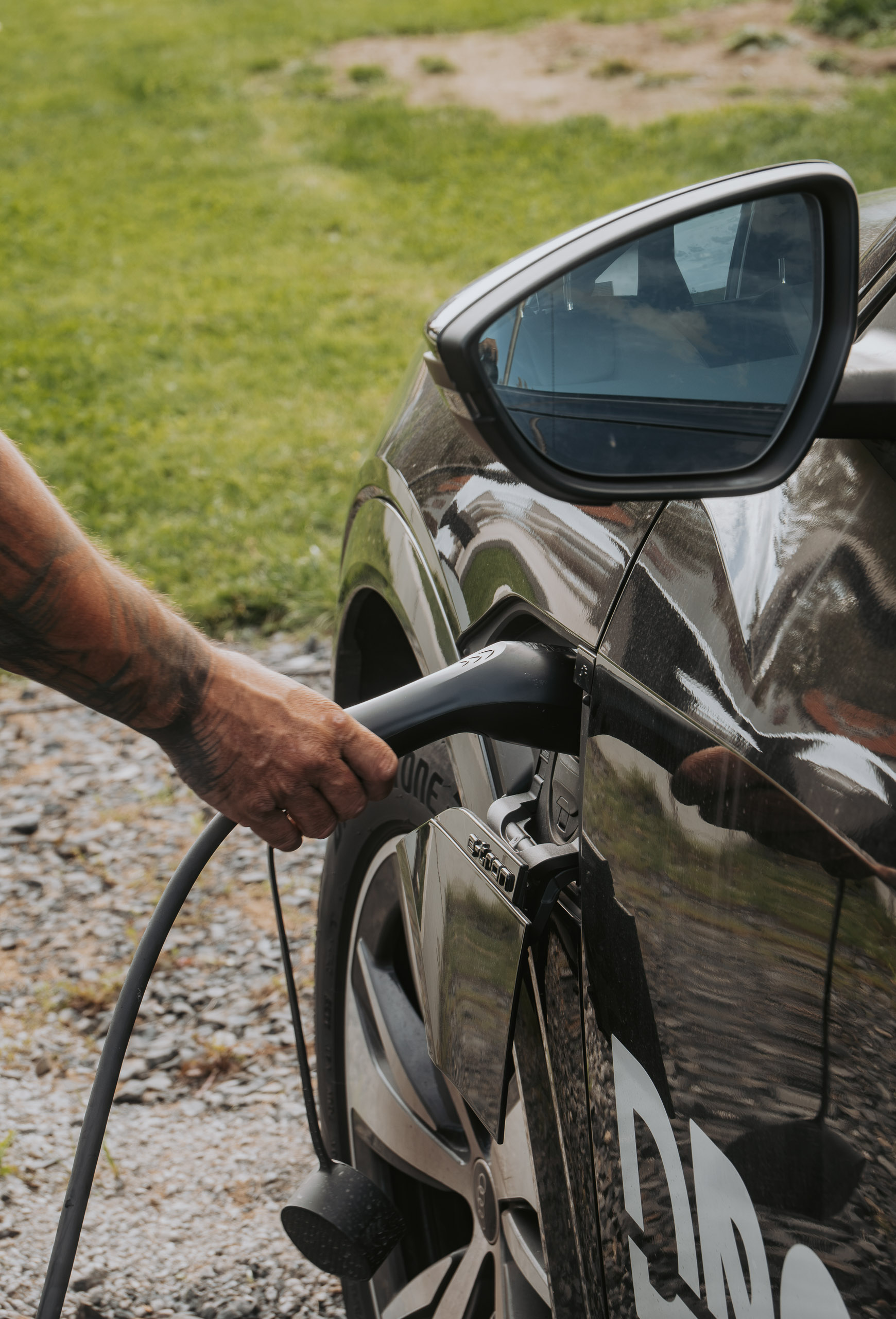 Eine Hand schließt ein Ladekabel an die Ladebuchse eines dunklen Elektroautos im Außenbereich an.