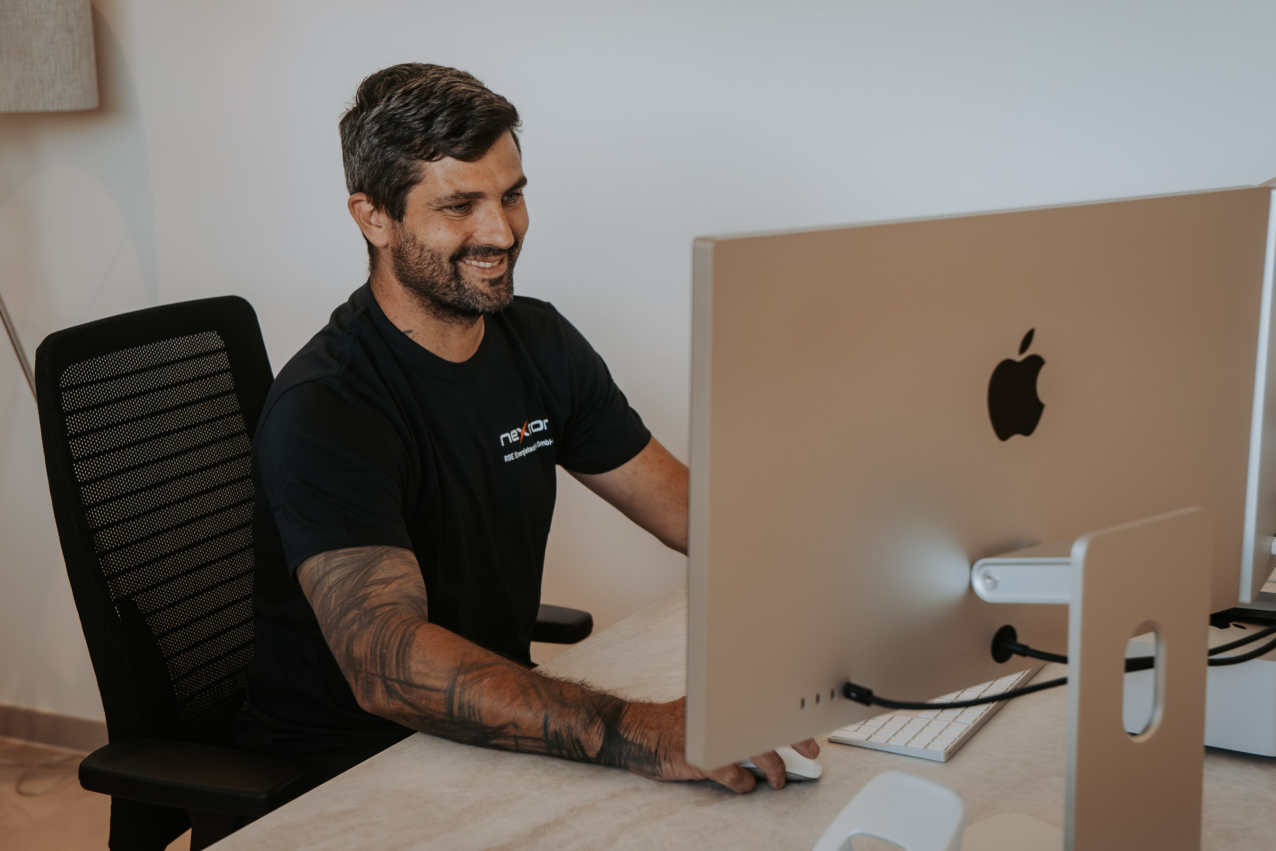 Ein lächelnder Mitarbeiter im schwarzen Nexron-T-Shirt arbeitet aufmerksam an einem Mac-Computer am Büro-Arbeitsplatz.
