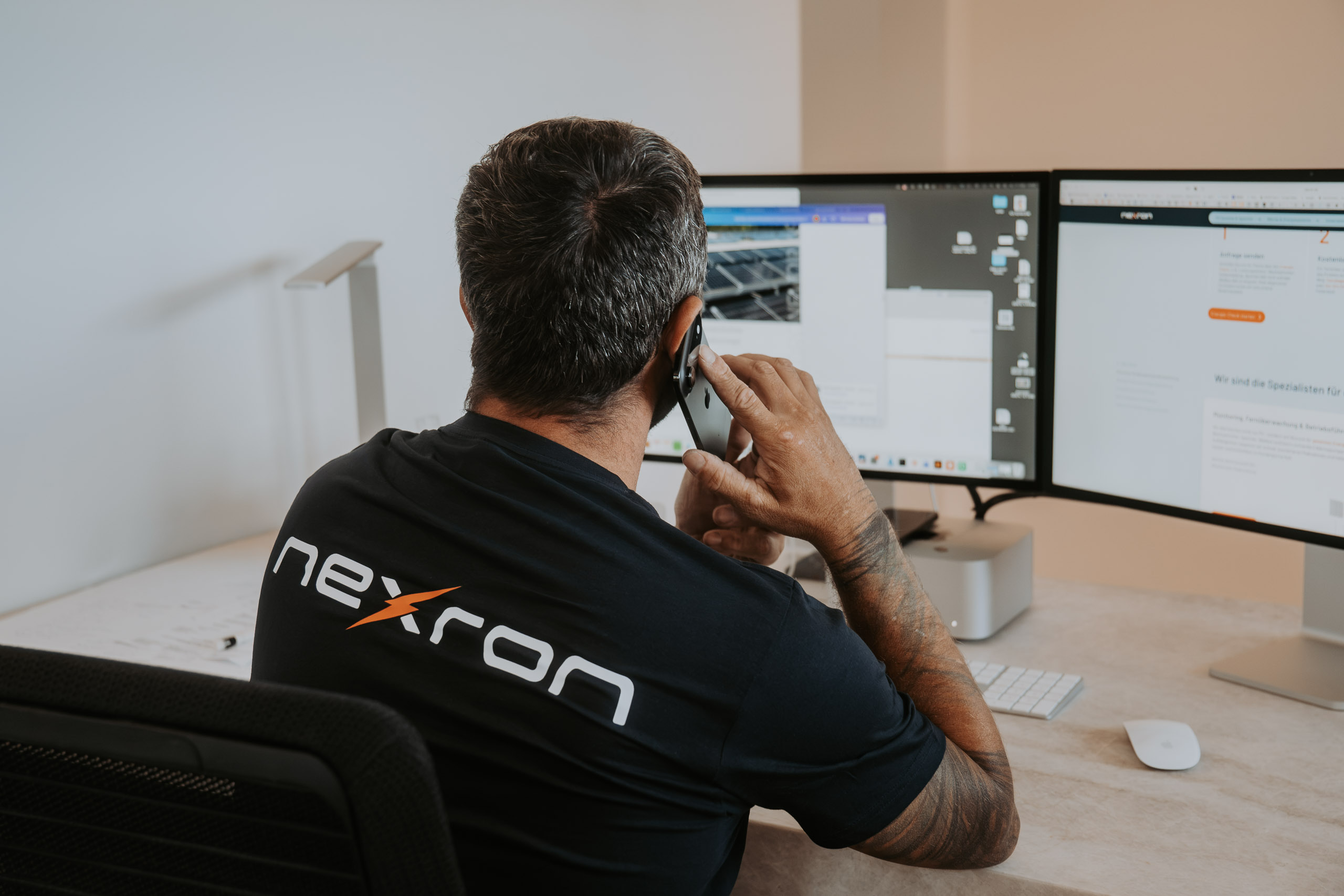 Ein Nexron-Mitarbeiter im schwarzen Firmen-T-Shirt telefoniert an einem Büro-Arbeitsplatz vor zwei Bildschirmen.