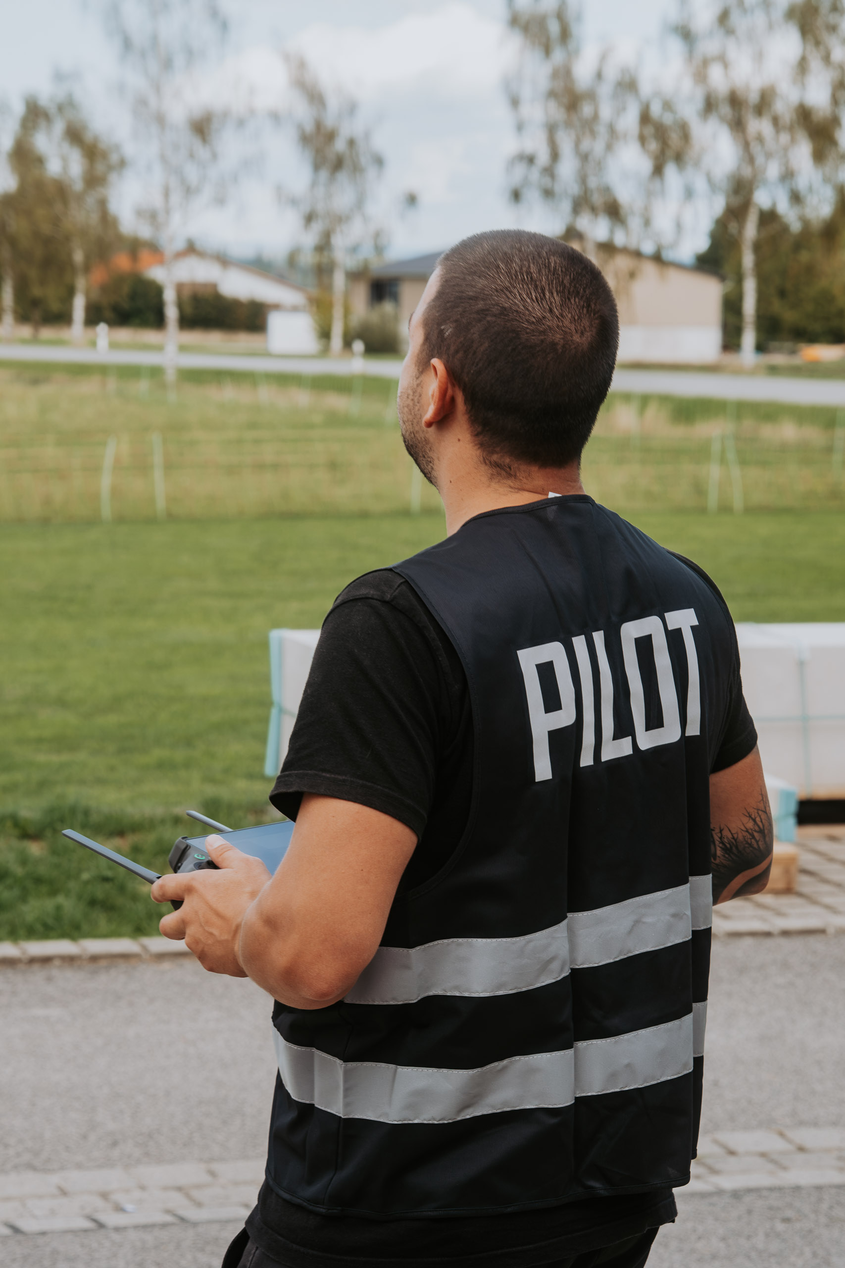 Person mit schwarzer Weste mit der Aufschrift „Pilot“ steuert eine Drohne im Freien.