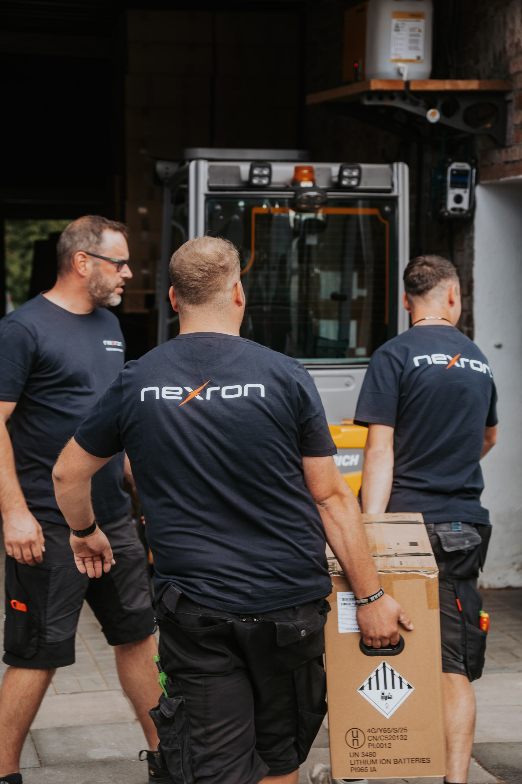 Drei Mitarbeiter der Nexron RSE Energietechnik GmbH tragen einen Karton mit Lithium-Ionen-Batterien im Lagerbereich vor einem Gabelstapler.