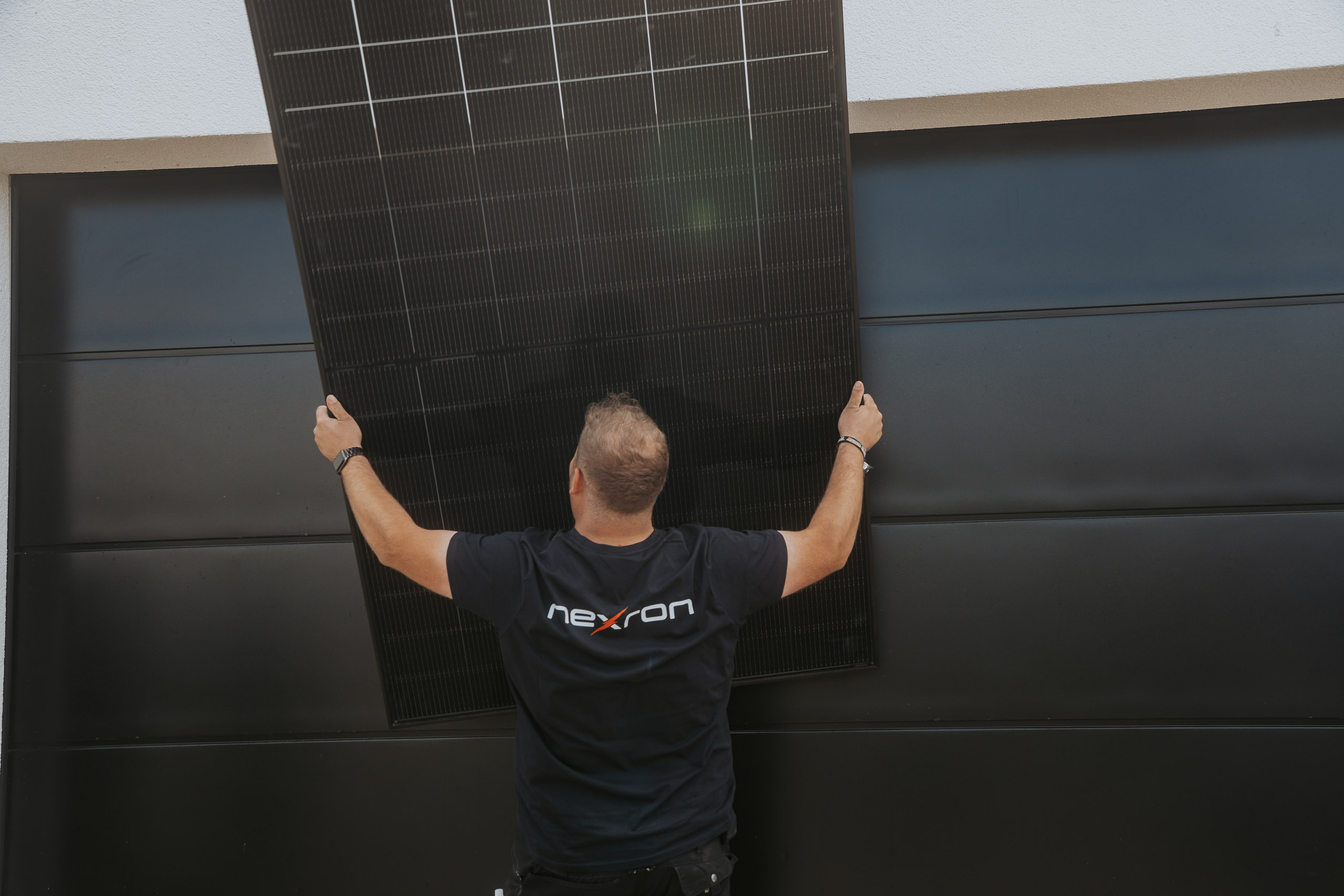 Ein Techniker im Nexron-Shirt hebt ein großes schwarzes Solarmodul vor einer dunklen Garagenfront empor.
