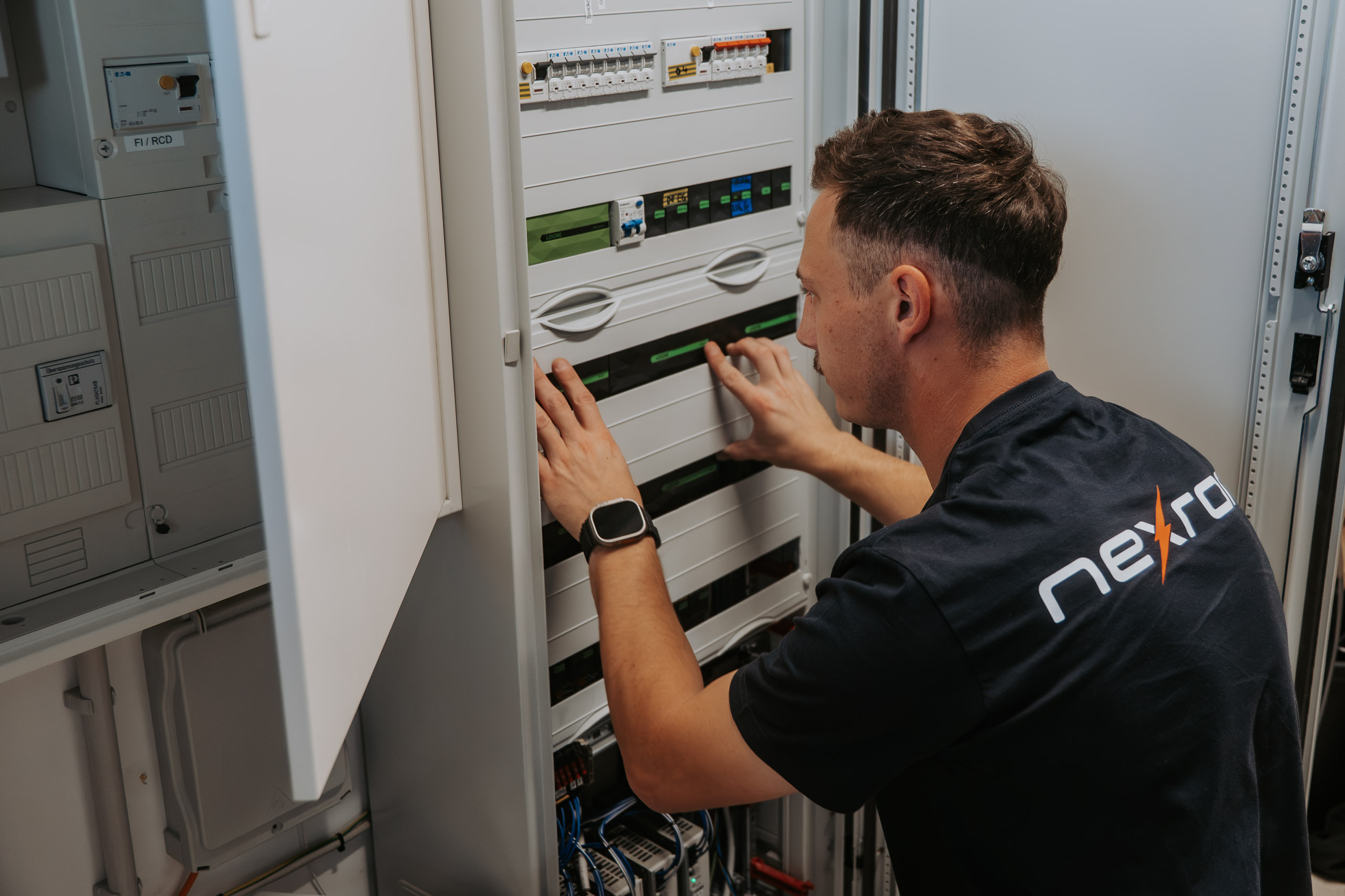 Ein Techniker im nexron-Shirt führt Arbeiten an den Sicherungen und Modulen eines geöffneten Schaltschranks durch.
