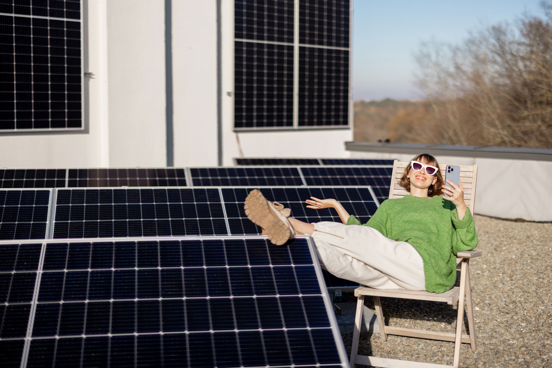 Frau im grünen Pullover entspannt auf Liegestuhl mit Smartphone neben Solarmodulen auf Hausdach.