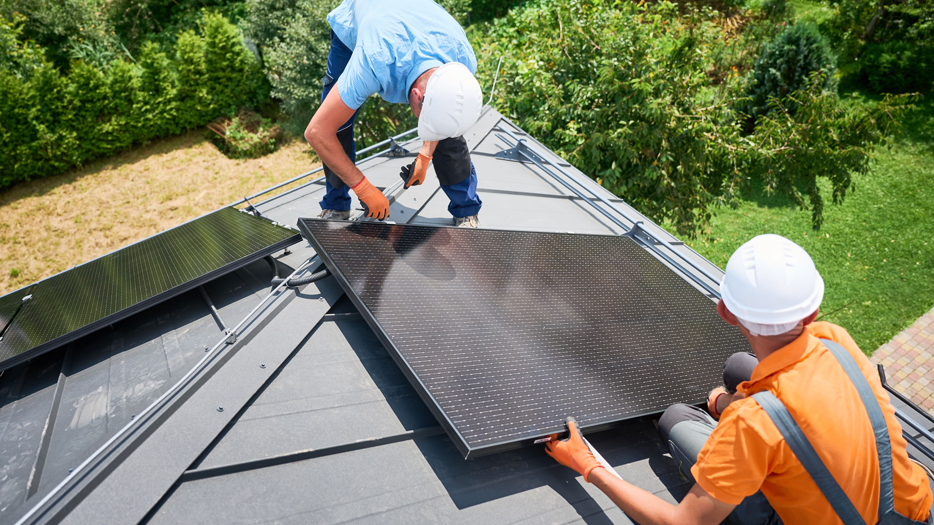 Zwei Servicetechniker mit Schutzhelmen arbeiten auf einem Hausdach an Solarmodulen und richten ein PV-Panel auf der Montageschiene aus.