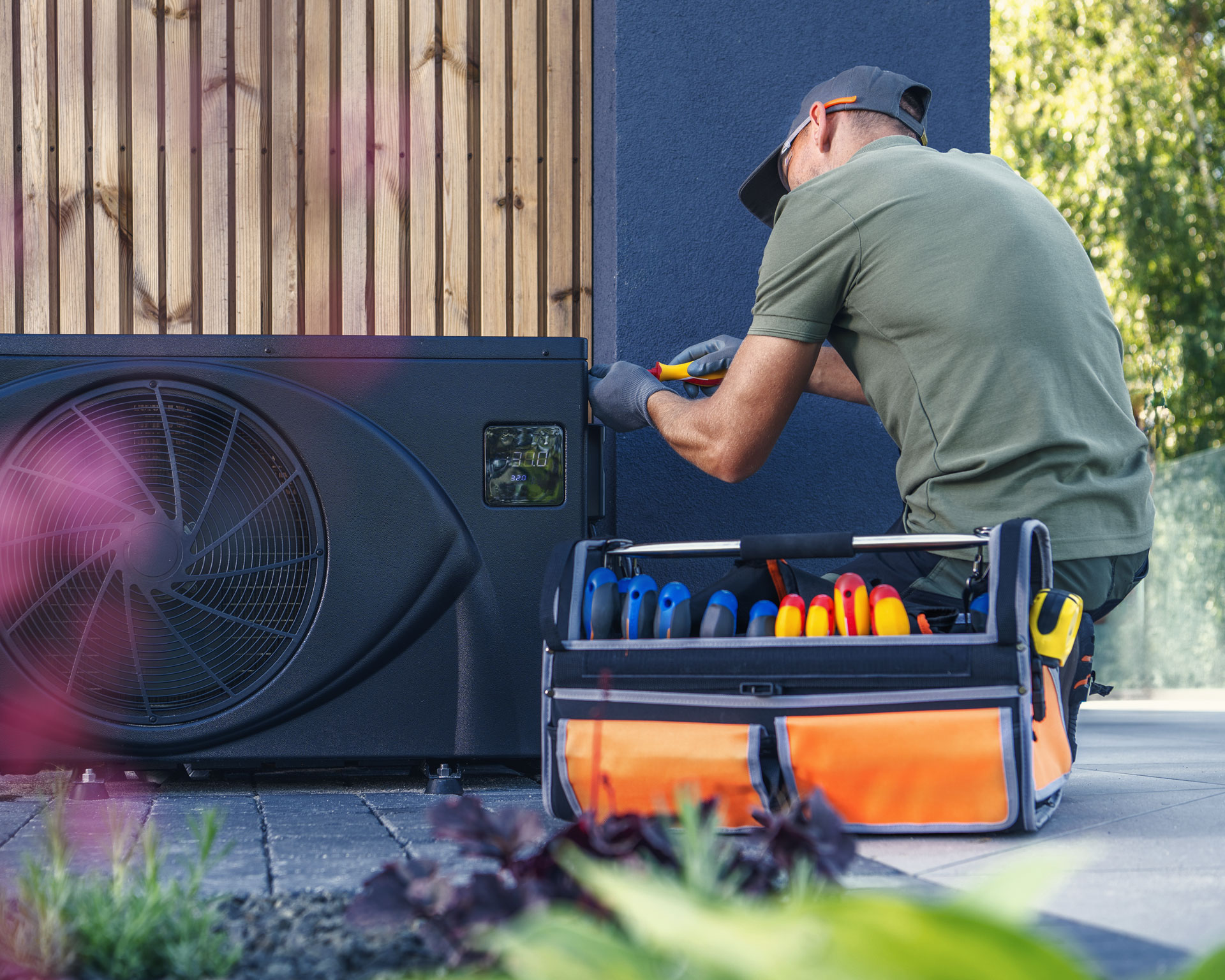 Techniker wartet eine Aussen-Waermepumpe vor einem Haus und arbeitet mit Werkzeug; daneben steht eine offene Werkzeugtasche.