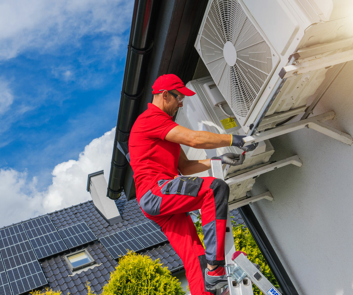 Monteur auf einer Leiter wartet das Aussengeraet einer Waermepumpe an der Hausfassade; im Hintergrund sind Solarmodule auf dem Dach zu sehen.